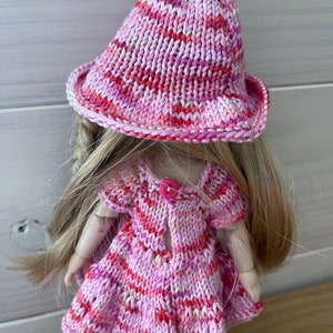 May include: A pink and white striped knitted dress and hat set for a doll. The dress has a button closure in the back and the hat has a wide brim.