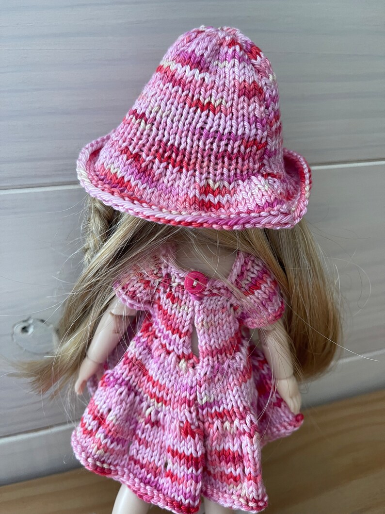 May include: A pink and white striped knitted dress and hat set for a doll. The dress has a button closure in the back and the hat has a wide brim.