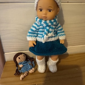 Peut inclure: Une poupée vêtue d'un pull, d'un chapeau et d'une jupe rayés bleu et blanc, avec des chaussettes blanches en tricot. La poupée a les cheveux bruns et les yeux bruns. Une plus petite poupée est couchée sur une surface en bois.