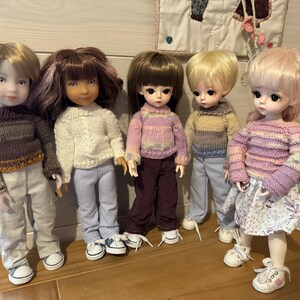 May include: Five dolls wearing knitted sweaters and pants. The dolls have different hair colors and styles. The dolls are standing on a wooden surface.