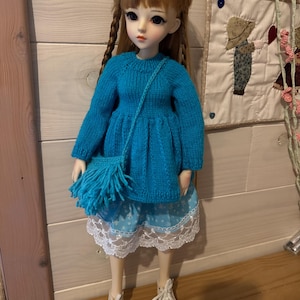 Peut inclure: Une poupée portant un pull en tricot bleu avec un sac à franges bleu assorti, une jupe bleu clair avec une bordure en dentelle blanche et des baskets blanches.