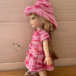 May include: A pink and white striped knitted dress and hat set for a doll. The dress has short sleeves and a flared skirt. The hat is a wide-brimmed sun hat.