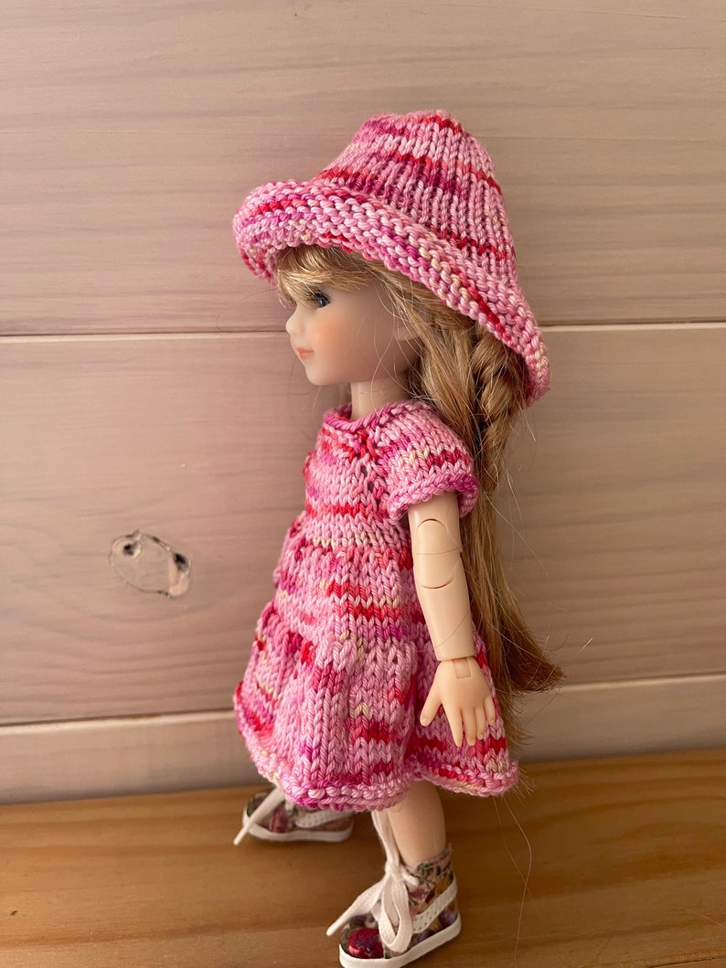 May include: A pink and white striped knitted dress and hat set for a doll. The dress has short sleeves and a flared skirt. The hat is a wide-brimmed sun hat.