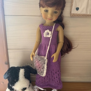 May include: A doll in a purple knitted dress with a white flower and a small crocheted purse. The doll has brown hair and is next to a black and white stuffed dog. The doll is wearing white trainers.