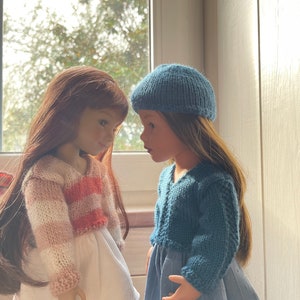 May include: Two dolls face each other, each with long hair. One doll wears a white dress and a striped pink and white sweater. The other doll wears a blue dress and a matching blue knitted cap and sweater. The dolls are positioned in front of a window.