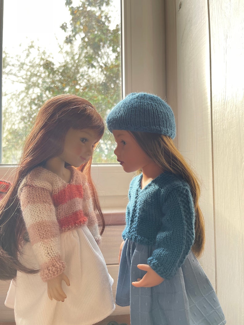 May include: Two dolls face each other, each with long hair. One doll wears a white dress and a striped pink and white sweater. The other doll wears a blue dress and a matching blue knitted cap and sweater. The dolls are positioned in front of a window.