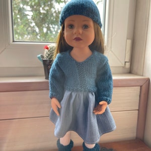 May include: A doll dressed in a knitted blue hat, sweater, and leg warmers, with a blue skirt and white socks. The doll has long brown hair and is positioned in front of a window.