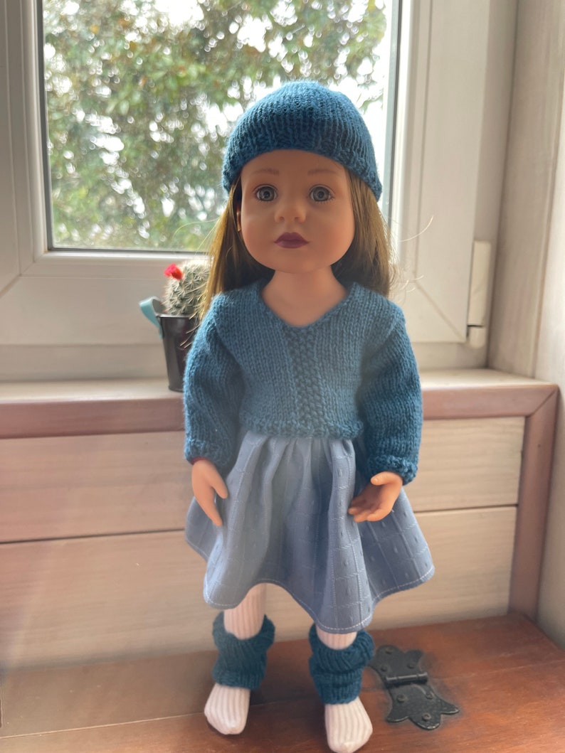 May include: A doll dressed in a knitted blue hat, sweater, and leg warmers, with a blue skirt and white socks. The doll has long brown hair and is positioned in front of a window.