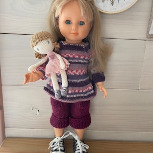 Peut inclure: Une poupée blonde aux yeux bleus, vêtue d'un pull et d'un pantalon en tricot violet. La poupée tient une petite poupée crochetée portant une robe rose. La poupée porte des baskets noires et blanches avec des lacets blancs. Un attrape-rêves est en arrière-plan.