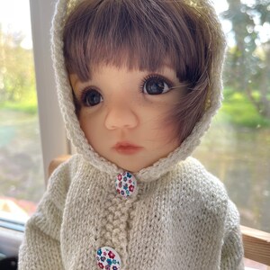 May include: A doll wearing a cream-colored knitted hooded sweater with floral buttons. The doll has brown hair and large, expressive eyes. The sweater appears handmade, with a textured knit pattern.