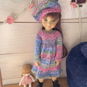 Puede incluir: Una muñeca con un vestido a rayas y una boina de punto de colores. La muñeca tiene el pelo castaño y los ojos verdes. La muñeca está de pie sobre una superficie de madera con una muñeca más pequeña sentada delante de ella.