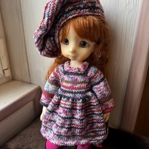 May include: A doll wearing a knitted outfit, including a beret and dress, in shades of pink, blue, and brown. The doll has red hair and is wearing pink shoes. The knitted dress has a round neckline and a flared skirt. The beret matches the dress.