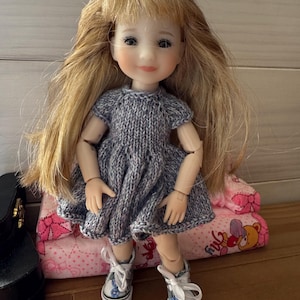 Peut inclure: Une poupée aux longs cheveux blonds portant une robe grise en tricot à manches courtes et des baskets blanches à lacets. La poupée possède des articulations aux épaules, aux coudes et aux poignets, ainsi que des traits peints, yeux et lèvres.