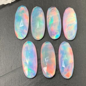 May include: Seven oval-shaped, faceted opals arranged in two rows. Each gemstone displays a translucent, milky base with iridescent flashes of pink, blue, green, and orange light reflecting across the polished, multi-faceted surfaces.