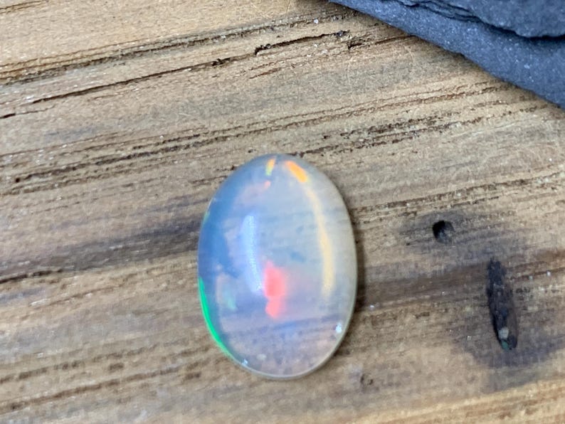 May include: Oval-shaped opal gemstone with a milky white base and flashes of green, orange, and red.