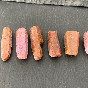 May include: Six raw pink and brown ruby crystals on a dark gray surface.