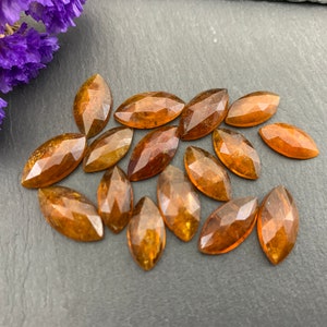 May include: Marquise-cut gemstones in amber and brown hues, scattered on a dark grey surface. The faceted gems reflect light, showcasing their polished surfaces. A sprig of purple flowers is in the upper left corner. The gems are suitable for jewellery making.