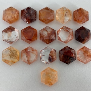 May include: A collection of sixteen hexagonal crystals in shades of orange, brown, and clear. The crystals are arranged in a grid pattern, showcasing their multifaceted surfaces and natural variations. The crystals are displayed on a white surface.