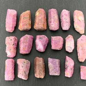 May include: A collection of raw ruby crystals, showcasing a range of pink and reddish hues. The gemstones vary in size and shape, with visible natural textures and dark inclusions. The crystals are arranged on a dark surface.