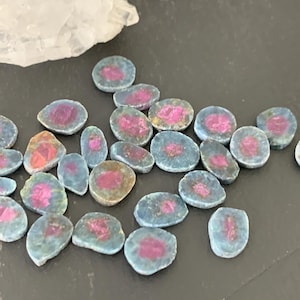 May include: A collection of polished, oval-shaped gemstones with a vibrant pink center surrounded by a blue-green hue. The stones vary slightly in size and shape, and are scattered across a dark surface. A piece of clear quartz is in the background.