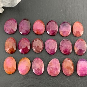 May include: A collection of oval-shaped ruby gemstones in various shades of red and pink. The faceted stones are arranged on a dark surface, showcasing their polished surfaces and natural color variations. Some stones have a deep, rich red hue, while others are lighter pink.