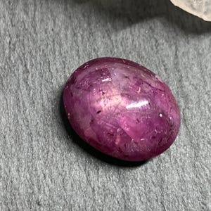 Op de afbeelding: Een gepolijste, ovaalvormige edelsteen in tinten van diep roze en magenta. De steen heeft een glad, reflecterend oppervlak met interne lichtspeling, tegen een grijze achtergrond met textuur. De edelsteen is waarschijnlijk een robijn of een vergelijkbare edelsteen.