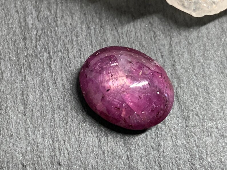 Op de afbeelding: Een gepolijste, ovaalvormige edelsteen in tinten van diep roze en magenta. De steen heeft een glad, reflecterend oppervlak met interne lichtspeling, tegen een grijze achtergrond met textuur. De edelsteen is waarschijnlijk een robijn of een vergelijkbare edelsteen.