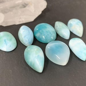 Peut inclure: Une collection de huit pierres pr&eacute;cieuses en cabochon bleues et blanches. Les pierres pr&eacute;cieuses sont de formes et de tailles vari&eacute;es, notamment en forme de larme et ovale. Les pierres pr&eacute;cieuses sont dispos&eacute;es sur une surface gris fonc&eacute;.