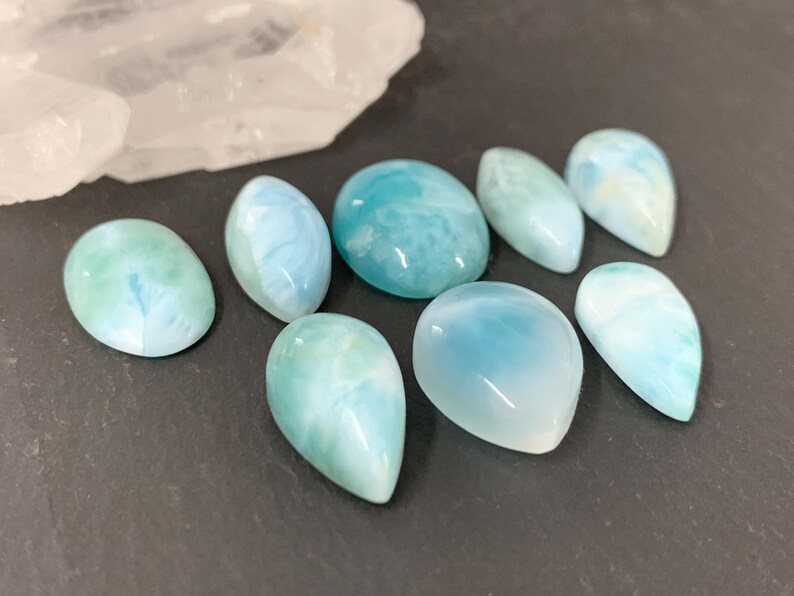 Peut inclure: Une collection de huit pierres pr&eacute;cieuses en cabochon bleues et blanches. Les pierres pr&eacute;cieuses sont de formes et de tailles vari&eacute;es, notamment en forme de larme et ovale. Les pierres pr&eacute;cieuses sont dispos&eacute;es sur une surface gris fonc&eacute;.