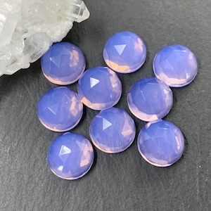 May include: A collection of nine faceted, round, light purple gemstones with a pinkish base. A cluster of white crystals is in the upper left corner. The gemstones are arranged on a dark gray surface.