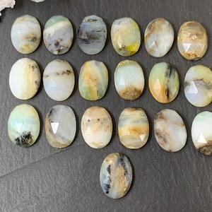 May include: A selection of oval gemstones with a faceted cut. The stones feature a range of colours, including white, yellow, green, and brown, with distinctive patterns and inclusions. The gemstones are displayed on a dark grey surface.