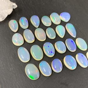 May include: A collection of 20 oval-shaped, iridescent opal gemstones. The opals are a milky white color with flashes of blue, green, and purple.