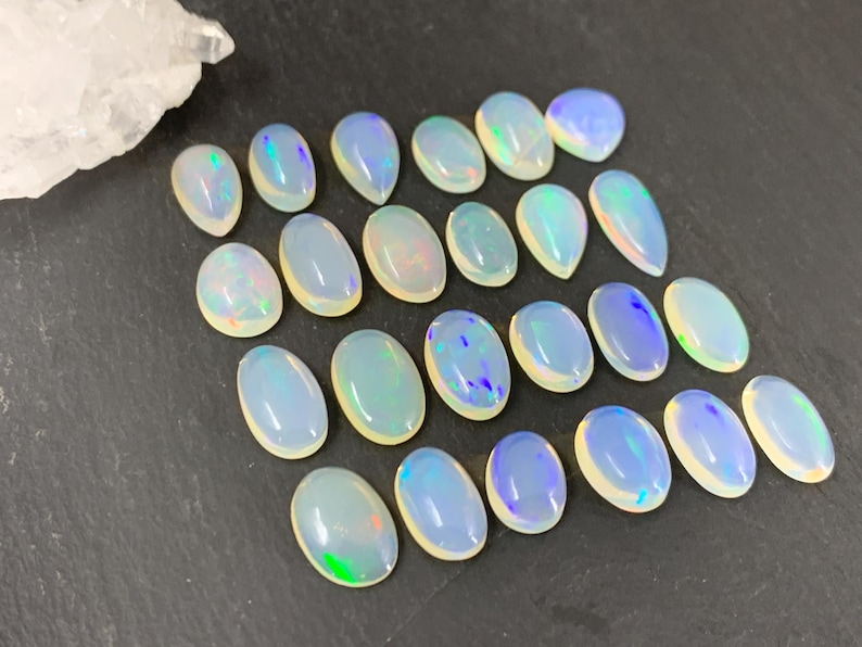 May include: A collection of 20 oval-shaped, iridescent opal gemstones. The opals are a milky white color with flashes of blue, green, and purple.