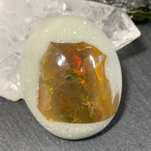 May include: Oval-shaped gemstone with a light speckled background. The center features a vibrant, iridescent opal with flashes of red, green, and gold. The opal is set within a speckled, off-white material, creating a striking contrast.