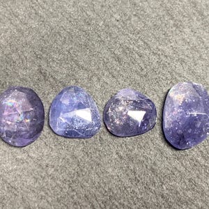 May include: Four faceted, polished gemstones in shades of purple and blue. The stones have irregular shapes, with a mix of oval, triangular, and rounded forms. The gems are displayed on a gray surface.