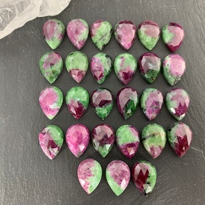 May include: A collection of 36 green and pink gemstone cabochons in a teardrop shape. The stones are arranged in rows on a dark gray surface.