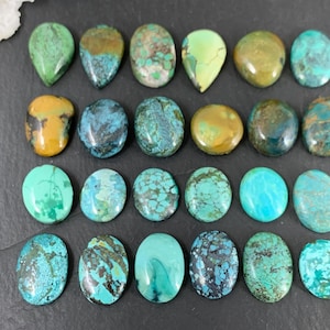 May include: A collection of twenty-one turquoise gemstones in various shapes, including oval, teardrop, and round. The stones display a range of colors from light to dark turquoise, with brown and black matrix patterns. The gemstones are arranged on a dark surface.