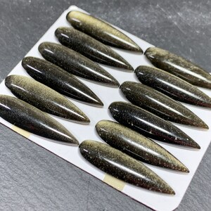 以下が含まれることがあります： 黒と金の宝石のカボションが12個セットで、それぞれが尖った楕円形をしています。カボションは白い背景に並べてあります。