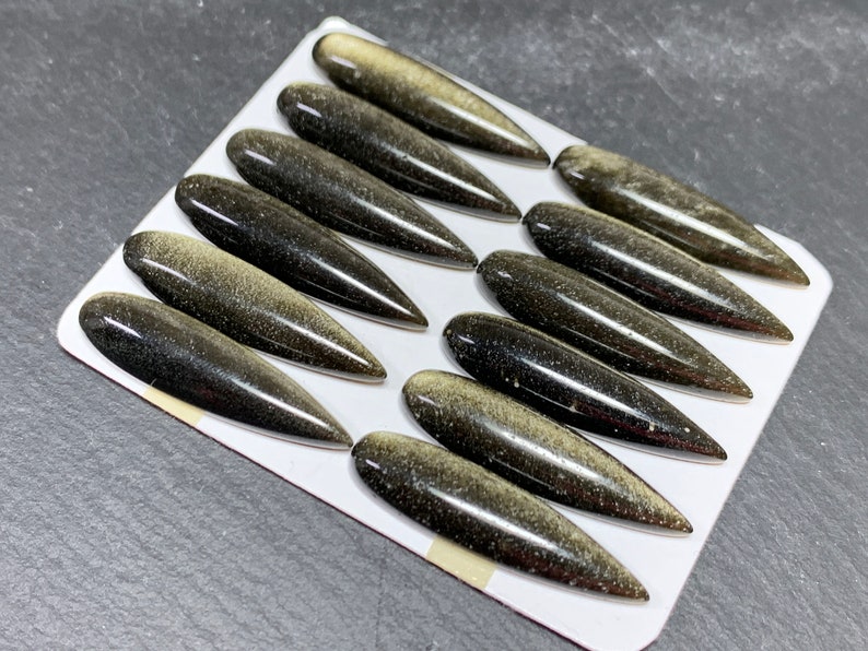 以下が含まれることがあります： 黒と金の宝石のカボションが12個セットで、それぞれが尖った楕円形をしています。カボションは白い背景に並べてあります。