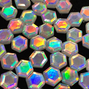 May include: A collection of iridescent, hexagonal-shaped beads. Each bead reflects a spectrum of colors, including blues, greens, yellows, and oranges, creating a shimmering effect. The beads are scattered against a dark background, highlighting their vibrant colors.
