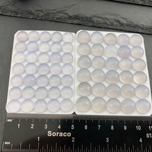 Op de afbeelding: Een verzameling lichtblauwe, ronde cabochons gerangschikt in twee witte trays. Een liniaal met inch- en millimeter maten is eronder geplaatst. Het woord "Soraco" is zichtbaar op de liniaal.