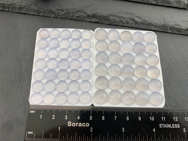 Op de afbeelding: Een verzameling lichtblauwe, ronde cabochons gerangschikt in twee witte trays. Een liniaal met inch- en millimeter maten is eronder geplaatst. Het woord "Soraco" is zichtbaar op de liniaal.