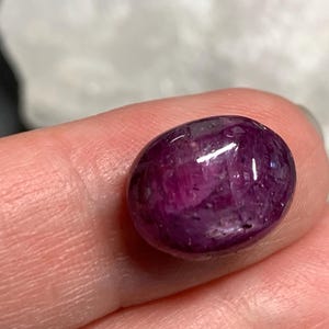 Op de afbeelding: Een gepolijste, ovaalvormige robijn edelsteen. De edelsteen heeft een diepe, verzadigde magenta kleur met een glanzend oppervlak. De steen wordt tussen twee vingers gehouden, met een vage achtergrond.