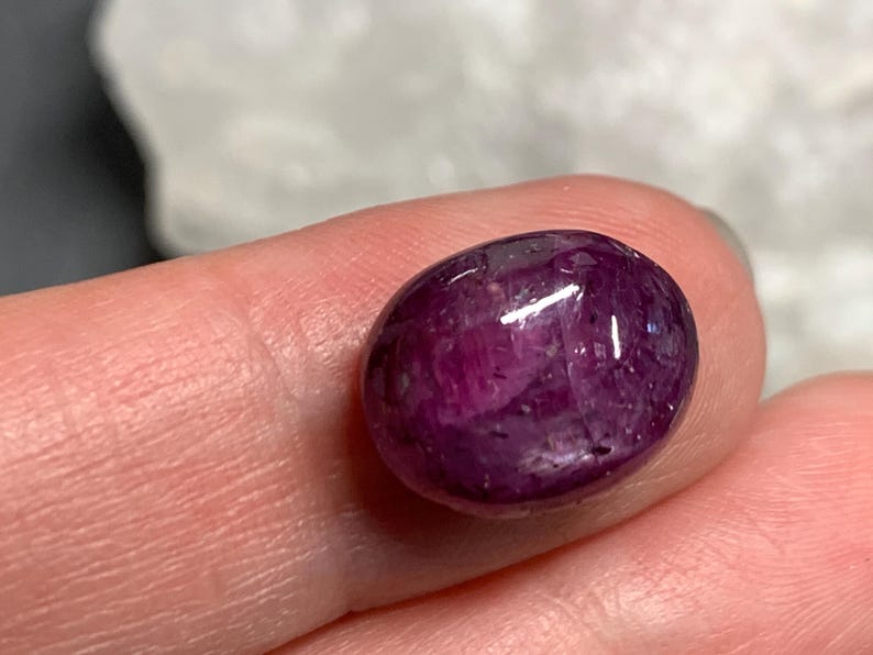 Op de afbeelding: Een gepolijste, ovaalvormige robijn edelsteen. De edelsteen heeft een diepe, verzadigde magenta kleur met een glanzend oppervlak. De steen wordt tussen twee vingers gehouden, met een vage achtergrond.