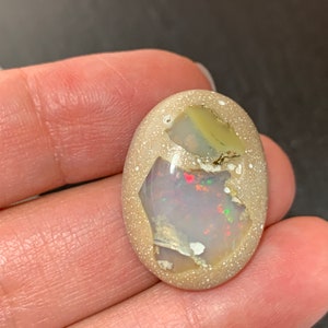 May include: Oval-shaped opal gemstone with a speckled, light tan exterior. The center reveals a vibrant display of colors, including red, green, and white. The opal is held in a hand, showcasing its natural beauty.