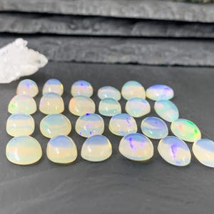 May include: A collection of 18 iridescent white opal cabochons, each with a slightly different pattern of color. The cabochons are arranged in a semi-circle on a dark gray surface.