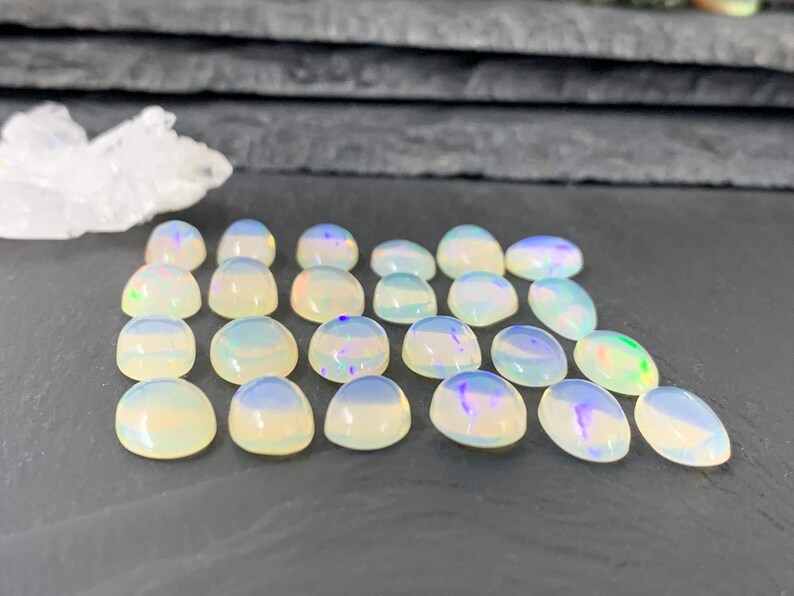 May include: A collection of 18 iridescent white opal cabochons, each with a slightly different pattern of color. The cabochons are arranged in a semi-circle on a dark gray surface.