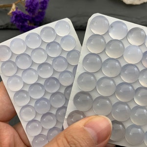 Op de afbeelding: Twee witte kaarten gevuld met talrijke kleine, ronde, lichtblauwe edelstenen. De edelstenen zijn gerangschikt in rijen en kolommen. De achtergrond omvat paarse bloemen en een donker oppervlak.