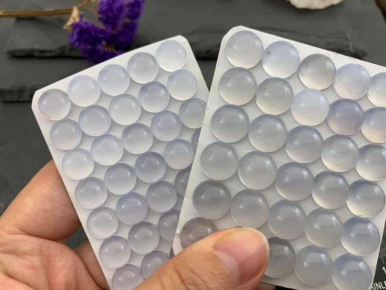 Op de afbeelding: Twee witte kaarten gevuld met talrijke kleine, ronde, lichtblauwe edelstenen. De edelstenen zijn gerangschikt in rijen en kolommen. De achtergrond omvat paarse bloemen en een donker oppervlak.