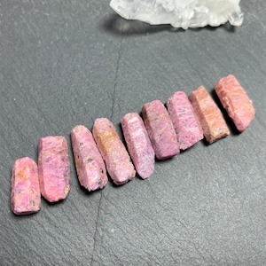 May include: A selection of raw ruby crystals, displaying a pinkish-red colour and a rough, textured surface. The crystals are arranged diagonally against a dark backdrop, with a clear quartz cluster visible in the background.